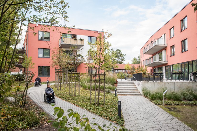 Ein modernes, rosafarbenes Wohnhaus mit barrierefreiem Zugang ist von gepflegtem Grün und jungen Bäumen umgeben. Zwei Personen im Rollstuhl bewegen sich über die Wege der Anlage. 
