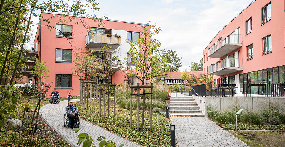 Ein modernes, rosafarbenes Wohnhaus mit barrierefreiem Zugang ist von gepflegtem Grün und jungen Bäumen umgeben. Zwei Personen im Rollstuhl bewegen sich über die Wege der Anlage.