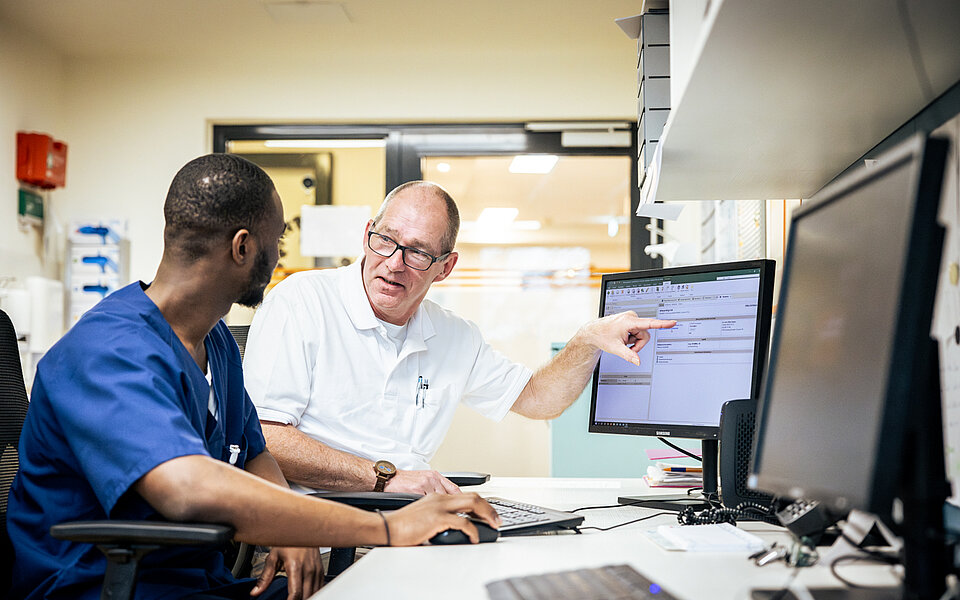 Ein älterer Mann in weißer Arbeitskleidung erklärt einem jüngeren Auszubildenden in blauer Arbeitskleidung etwas an einem Computerbildschirm in einem Arbeitsraum. 
