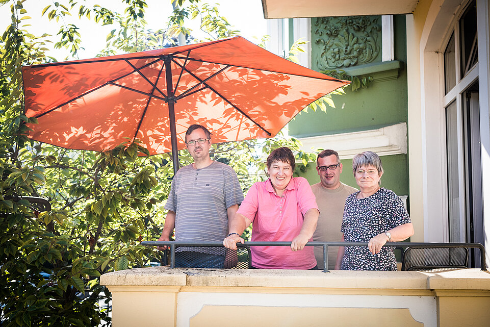 Vier Personen stehen auf einem sonnigen Balkon unter einem großen roten Sonnenschirm. Sie blicken freundlich in die Kamera. 