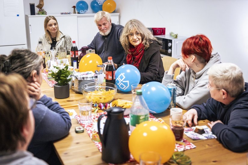 Mehrere Personen verschiedener Generationen sitzen gemeinsam an einem Tisch mit Snacks und Luftballons und unterhalten sich in gemütlicher Atmosphäre.