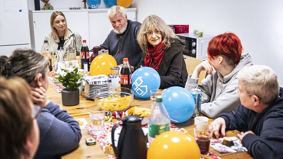 Mehrere Personen verschiedener Generationen sitzen gemeinsam an einem Tisch mit Snacks und Luftballons und unterhalten sich in gemütlicher Atmosphäre.