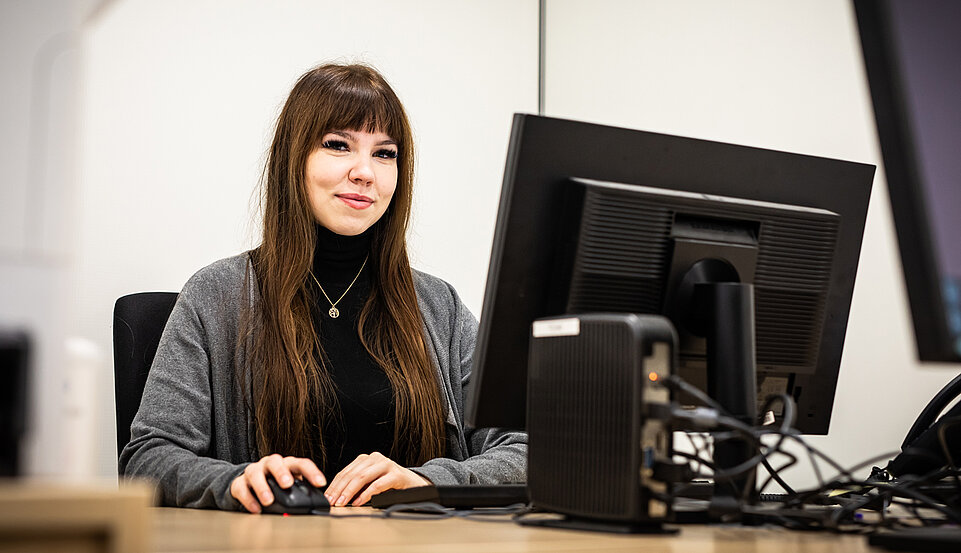 Eine junge Frau sitzt lächelnd an einem Schreibtisch und arbeitet an einem Computer.
