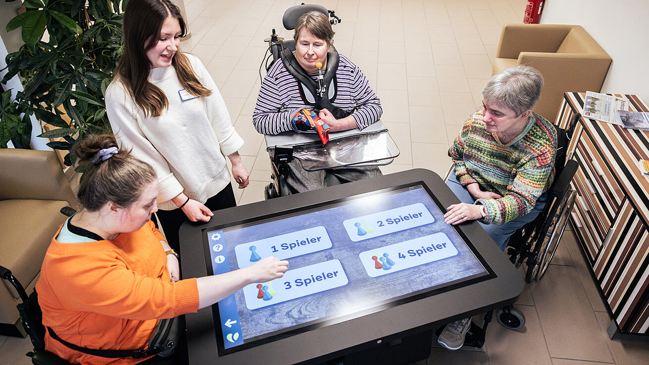 Vier Frauen spielen ein Spiel an einem übergroßen Tablet, das zwischen ihnen steht. Drei Frauen sitzen im Rollstuhl.