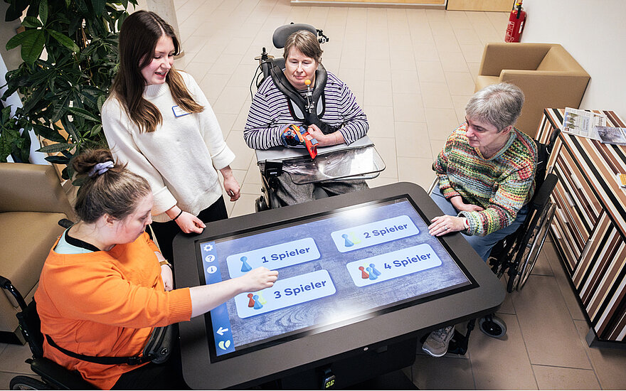 Vier Frauen spielen ein Spiel an einem übergroßen Tablet, das zwischen ihnen steht. Drei Frauen sitzen im Rollstuhl.