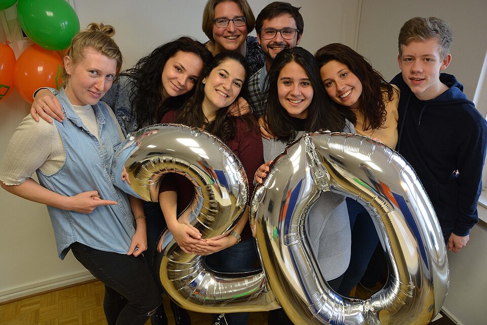 Acht Personen stehen eng beieinander und blicken in die Kamera. Vor ihnen halten sie silberne Luftballons in Form der Zahl „20“, im Hintergrund sind weitere bunte Luftballons zu sehen.