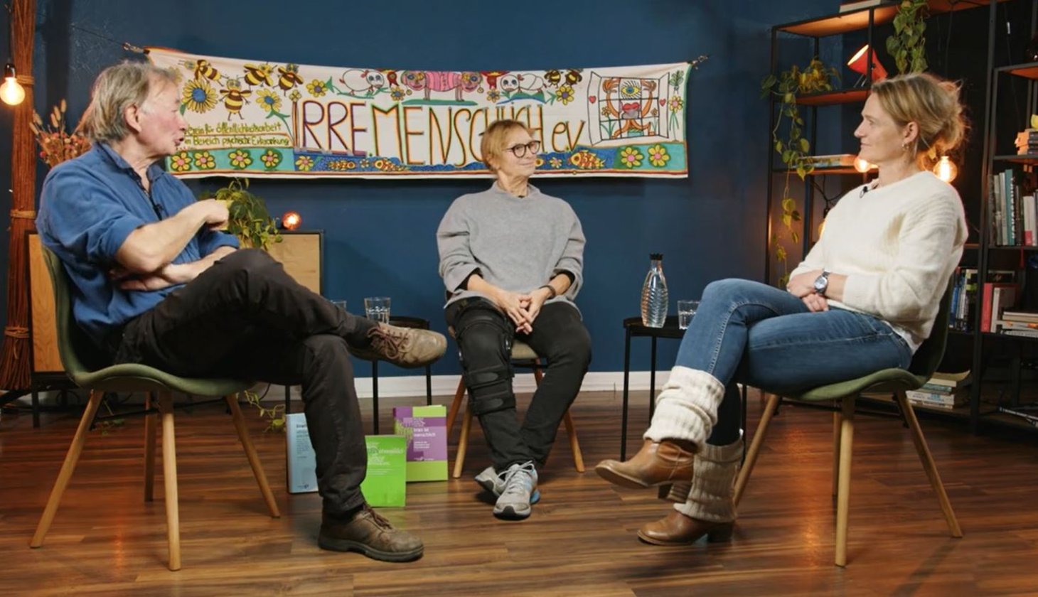 Drei Personen in Freizeitkleidung unterhalten sich und diskutieren, sitzen auf Stühlen in einem Raum vor einem Banner mit der Aufschrift „Irre Menschlich e.v.“ im Hintergrund.
