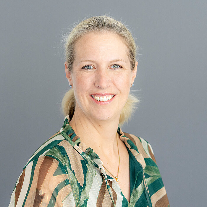 Porträt einer Person (Yvonne Stresow) mit zurückgebundenen blonden Haaren vor einem grauen Hintergrund. Die Person trägt ein gemustertes Oberteil unter einem dunklen Blazer und blickt in die Kamera.