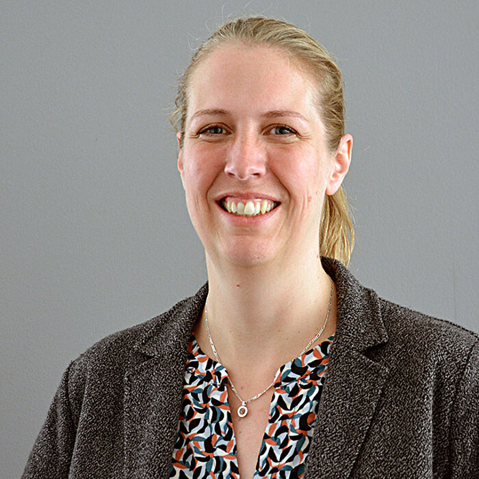 Porträt einer Person (Yvonne Stresow) mit zurückgebundenen blonden Haaren vor einem grauen Hintergrund. Die Person trägt ein gemustertes Oberteil unter einem dunklen Blazer und blickt in die Kamera.