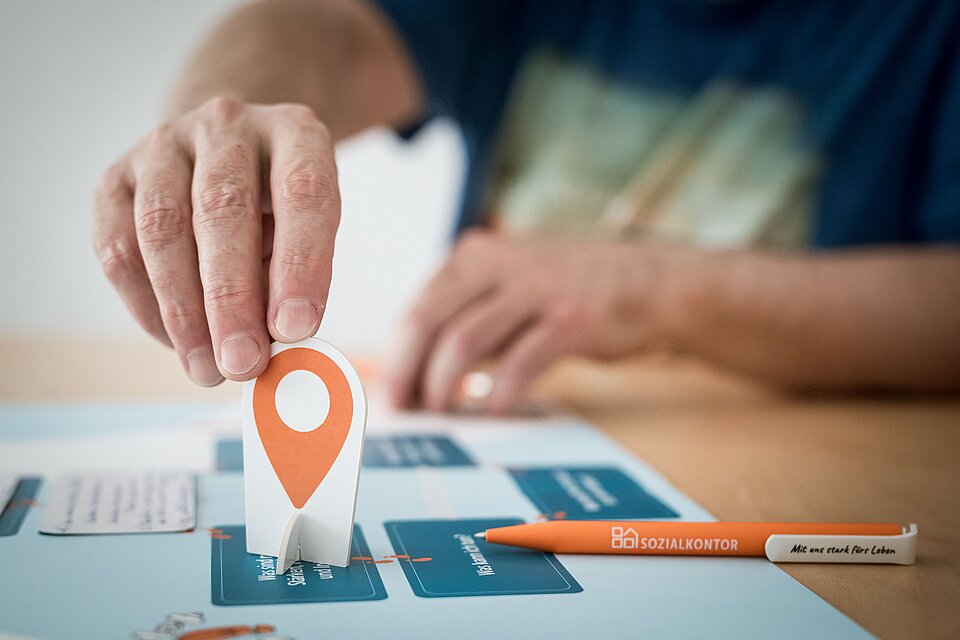 Eine Person platziert während einer Planungssitzung eine orangene Standortmarkierung auf einem blauem Plan mit Fragen. Daneben liegt ein Stift mit der Aufschrift 