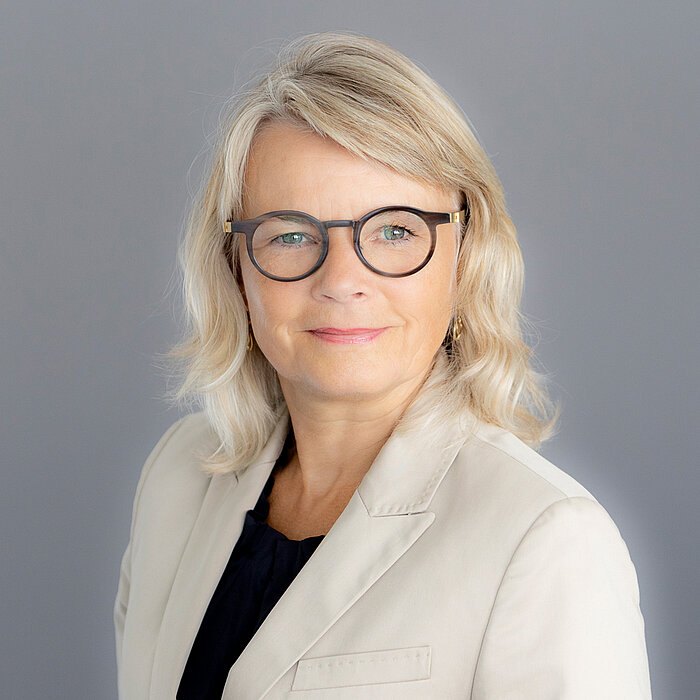 Nicola Pantelias, eine Frau mit lockigem, blondem Haar und Brille, lächelt freundlich in die Kamera. Sie trägt ein dunkles Oberteil. Der Hintergrund ist dunkelgrau gestaltet.