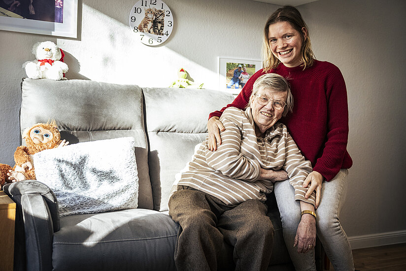Eine jüngere Frau in rotem Pullover sitzt auf einem grauen Sofa und umarmt eine ältere Frau mit kurzen Haaren und gestreiftem Pullover. Beide lächeln in die Kamera. Im Hintergrund stehen Kuscheltiere und an der Wand hängen Familienfotos und eine Uhr mit Katzenmotiv.