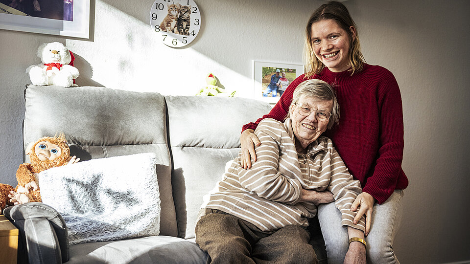 Eine jüngere Frau in rotem Pullover sitzt auf einem grauen Sofa und umarmt eine ältere Frau mit kurzen Haaren und gestreiftem Pullover. Beide lächeln in die Kamera. Im Hintergrund stehen Kuscheltiere und an der Wand hängen Familienfotos und eine Uhr mit Katzenmotiv.