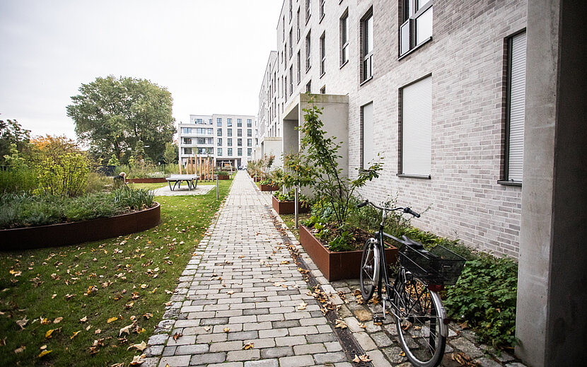 Ein moderner Wohnkomplex mit heller Fassade und barrierefreiem, gepflastertem Weg führt durch eine gepflegte Grünanlage. Links im Bild sind Hochbeete und Sitzgelegenheiten zu sehen.