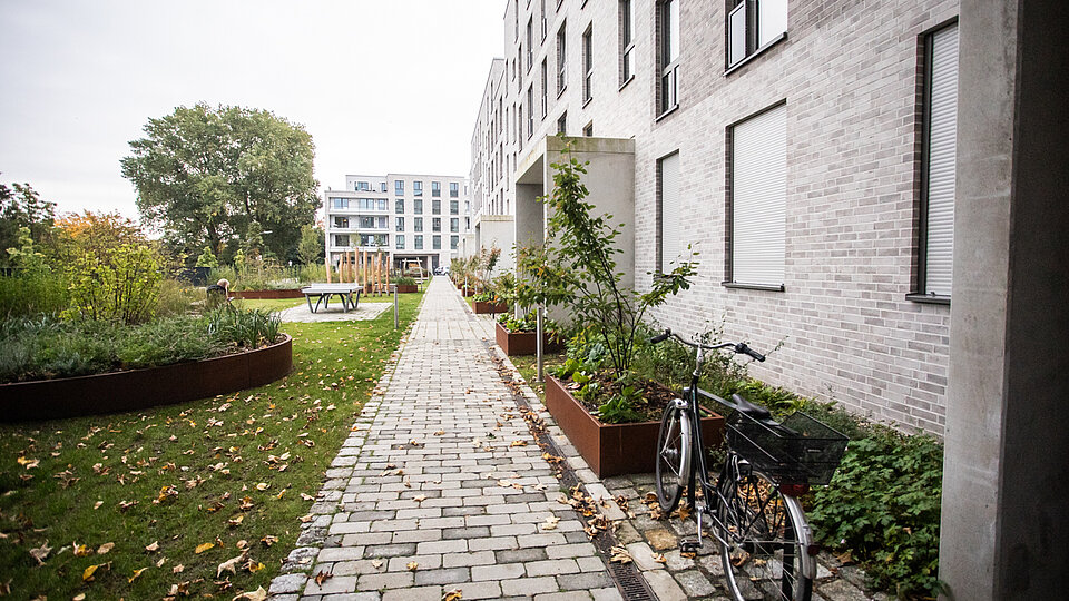 Ein moderner Wohnkomplex mit heller Fassade und barrierefreiem, gepflastertem Weg führt durch eine gepflegte Grünanlage. Links im Bild sind Hochbeete und Sitzgelegenheiten zu sehen.