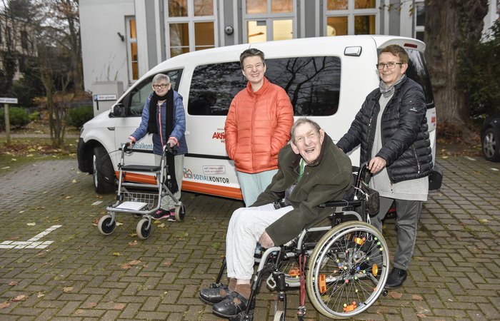 Vier Personen, davon eine im Rollstuhl vor einem Auto stehend