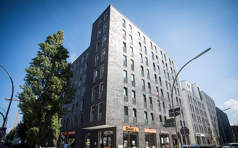 Ein großes, modernes Wohn- und Geschäftshaus mit dunkler Fassade erstreckt sich über mehrere Stockwerke an einer belebten Straße. Im Erdgeschoss befinden sich Schaufenster und Eingänge.