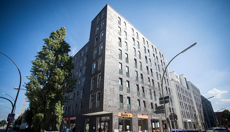 Ein großes, modernes Wohn- und Geschäftshaus mit dunkler Fassade erstreckt sich über mehrere Stockwerke an einer belebten Straße. Im Erdgeschoss befinden sich Schaufenster und Eingänge.