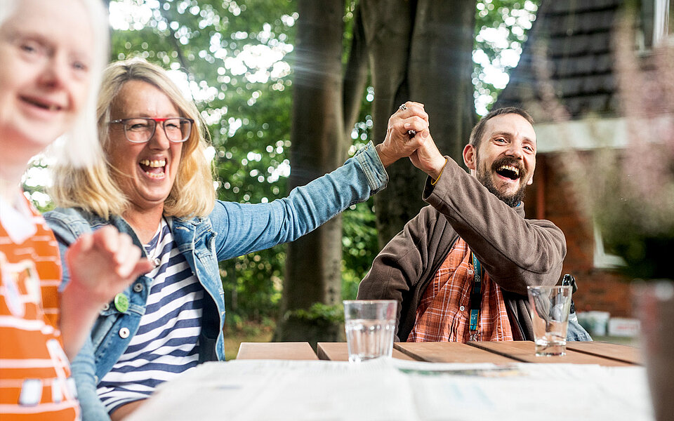 Zwei Personen ringen spielerisch an einem Tisch im Freien mit den Armen und Händen und zeigen dabei Freude und Lachen. Links ist eine weitere Person zu sehen, die lächelt und den Moment mit ihnen genießt.