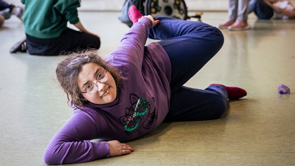 Eine Schülerin liegt lachend auf dem Boden einer Turnhalle und führt eine Bewegung aus. Im Hintergrund sind weitere Teilnehmende des Hip-Hop-Projekts zu sehen.