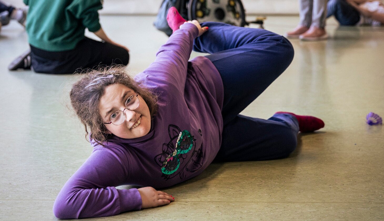Eine Schülerin liegt lachend auf dem Boden einer Turnhalle und führt eine Bewegung aus. Im Hintergrund sind weitere Teilnehmende des Hip-Hop-Projekts zu sehen.