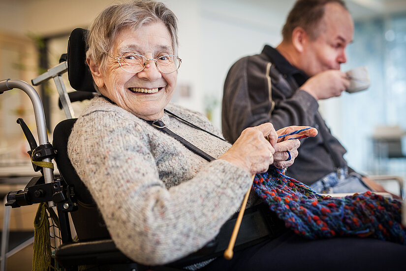 Ältere Frau im Rollstuhl strickt, Mann trinkt aus Tasse