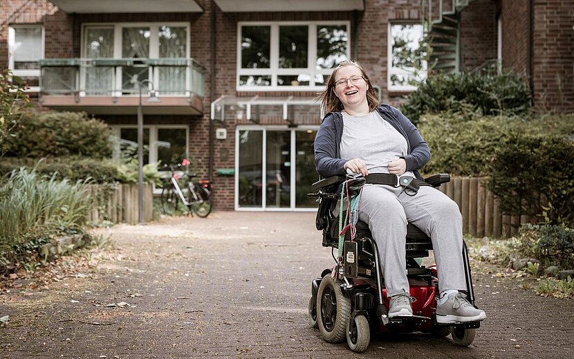 Eine Frau im Elektrorollstuhl sitzt lächelnd vor dem Eingang eines modernen Wohngebäudes mit Balkonen. Der Weg ist barrierefrei gestaltet.