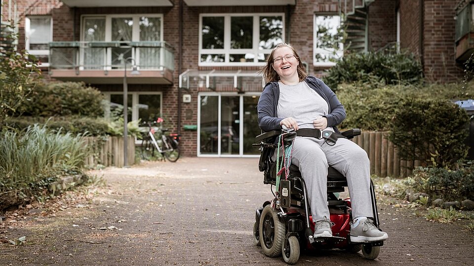 Eine Frau im Elektrorollstuhl sitzt lächelnd vor dem Eingang eines modernen Wohngebäudes mit Balkonen. Der Weg ist barrierefrei gestaltet.