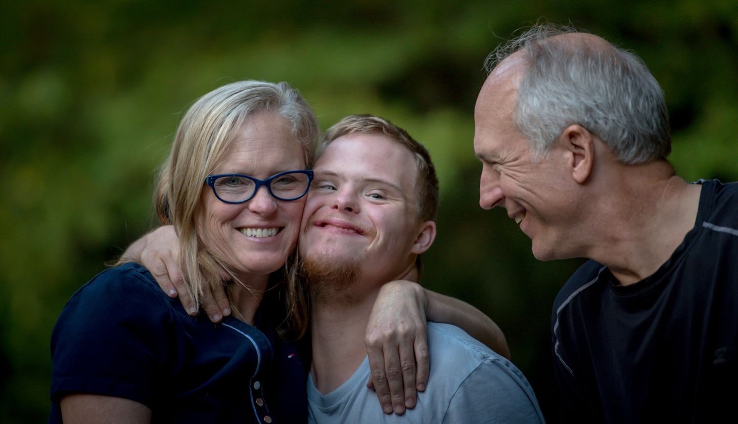 Zwei erwachsene Personen umarmen liebevoll eine jüngere Person mit Down-Syndrom. Alle lächeln.
