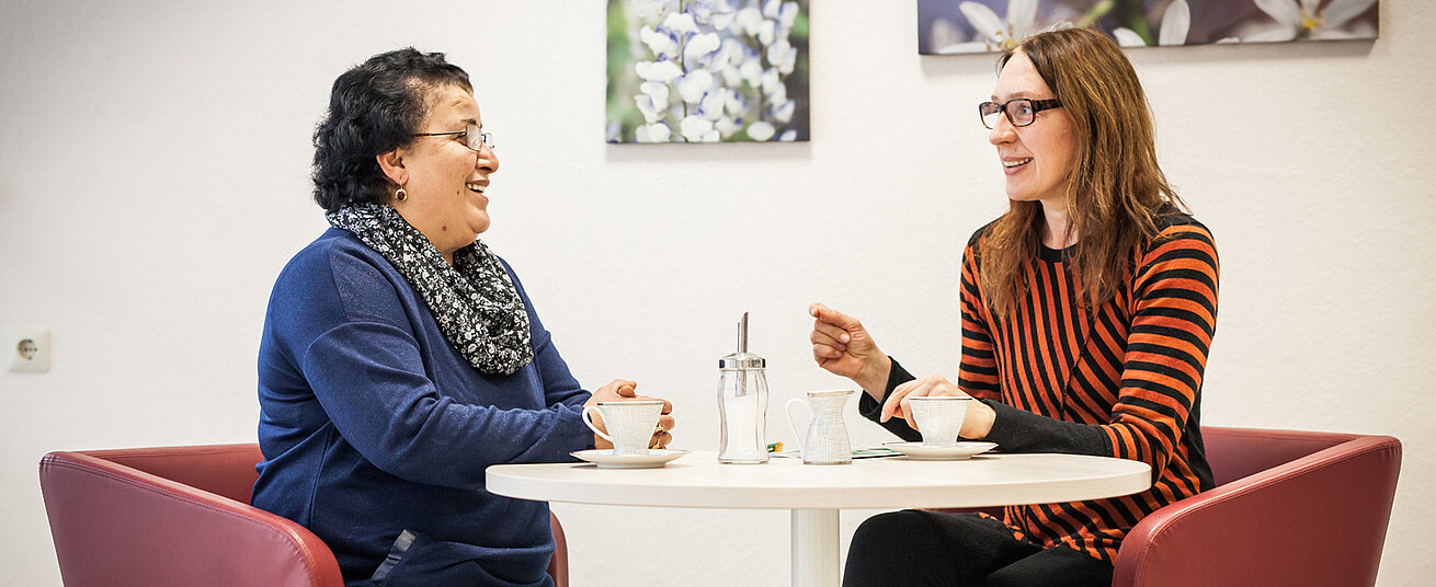 In einem Raum. Zwei Frauen sitzen, sich unterhaltend, an einem runden Tisch gegenüber.