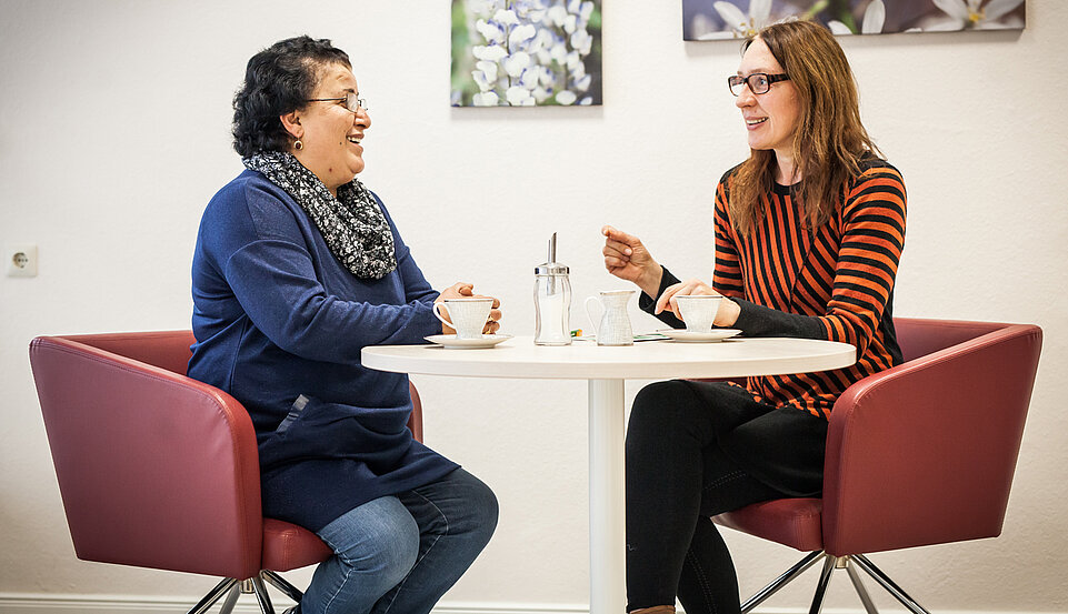 In einem Raum. Zwei Frauen sitzen, sich unterhaltend, an einem runden Tisch gegenüber.